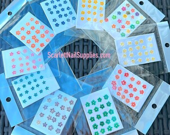 May include: Ten individual packages of nail art stickers featuring small flower designs in various colors, including blue, yellow, pink, orange, green, and purple. The packages are labeled with the website "ScarlettNailSupplies.com".