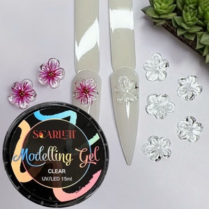 May include: A clear modeling gel in a black jar with the brand name "SCARLETT" and the text "Modelling Gel CLEAR UV/LED 15ml". The jar is surrounded by small, clear and pink flower-shaped nail decorations.