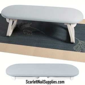May include: A white foldable nail table with a light gray padded surface. The table is set up on a black and beige mat with a floral design. The table is folded in the second image. The text "ScarlettNailSupplies.com" is visible at the bottom of the image.