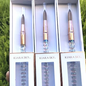 May include: Three nail art brushes with iridescent handles and gold accents. The brushes are packaged in white boxes with the brand name "KIARA SKY" printed on the front.