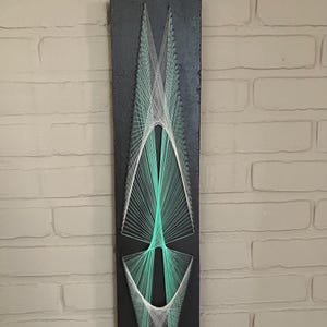 Könnte beinhalten: Ein vertikales, rechteckiges schwarzes Holzbrett mit Fadenkunst aus mintgrünem und weißem Garn. Die Fäden sind um kleine Nägel gewickelt und bilden zwei symmetrische, längliche geometrische Formen mit spitzen Enden und offenen Zentren.