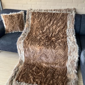 Op de afbeelding: Een bruine faux fur plaid en bijpassende kussenset. De deken heeft een visgraatpatroon en een lichtbruine rand. Het kussen is vierkant en heeft een vergelijkbare textuur. De set wordt gepresenteerd op een donkergrijze bank.