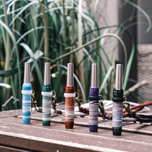 May include: Five cylindrical whistles with metallic silver tops and marbled resin bodies in blue, green, rust, purple, and black. Each whistle features a white or black silicone ring and a metal clasp attached to a braided cord lanyard.