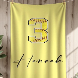 May include: A yellow softball towel with the number 3 in a softball design. The number is outlined in black with red stitching. The name "Hannah" is written in a cursive font below the number.
