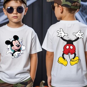 May include: A white short-sleeve t-shirt featuring a graphic of Mickey Mouse's head peeking through a torn fabric effect on the front, and a graphic of Mickey Mouse's lower body and legs on the back, showing red shorts and yellow shoes.
