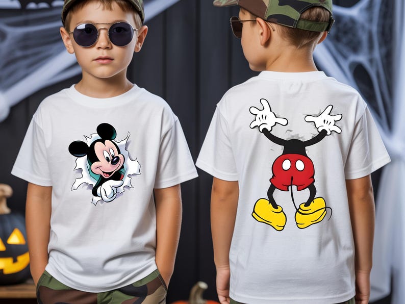 May include: A white short-sleeve t-shirt featuring a graphic of Mickey Mouse's head peeking through a torn fabric effect on the front, and a graphic of Mickey Mouse's lower body and legs on the back, showing red shorts and yellow shoes.