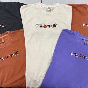 Puede incluir: Cinco camisetas coloridas en tonos de gris, crema y naranja, cada una bordada con un diseño temático de Halloween. Los diseños incluyen un fantasma, un sombrero de bruja, una calabaza, un murciélago y una lápida. Las camisetas son de un material suave y cómodo.