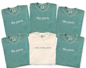 Puede incluir: Cinco camisetas de color verde azulado con el texto blanco the party impreso en el pecho y una camiseta de color crema con el texto verde azulado wife of the party impreso en el pecho. Cada camiseta tiene cuello redondo y etiqueta de marca.