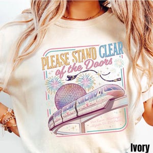 Puede incluir: Camiseta de color marfil con un gr&aacute;fico colorido que presenta un monorra&iacute;l, fuegos artificiales y el texto "PLEASE STAND CLEAR of the Doors". El dise&ntilde;o incluye una esfera y el texto "Por Favor, Mant&eacute;ngase Alejado De Las Puertas".