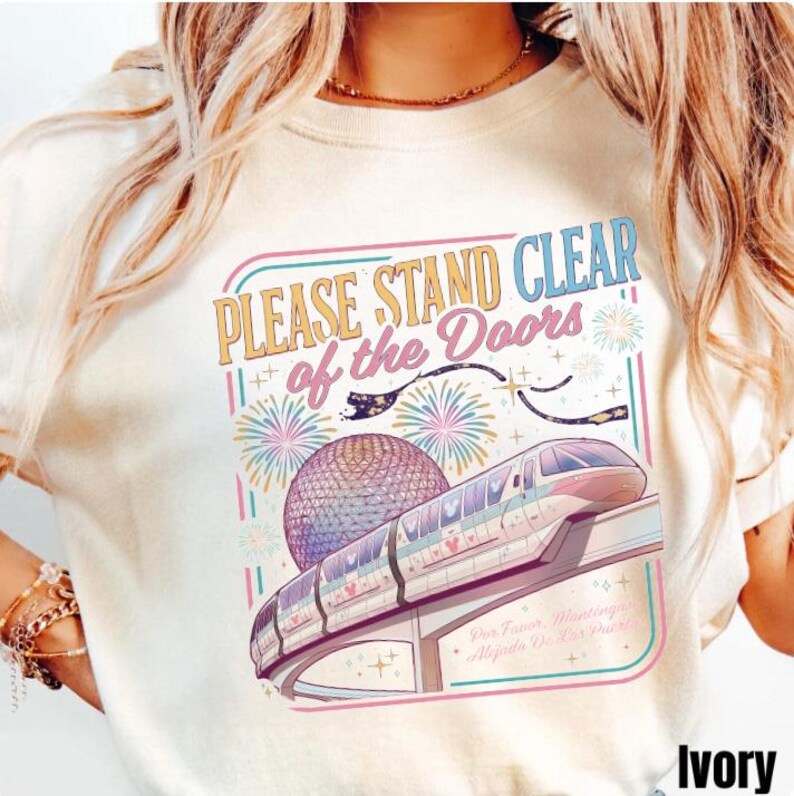 Puede incluir: Camiseta de color marfil con un gr&aacute;fico colorido que presenta un monorra&iacute;l, fuegos artificiales y el texto "PLEASE STAND CLEAR of the Doors". El dise&ntilde;o incluye una esfera y el texto "Por Favor, Mant&eacute;ngase Alejado De Las Puertas".