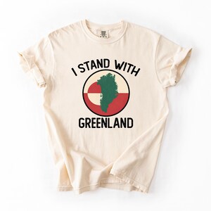 Pu&ograve; includere: T-shirt color crema con la scritta nera "I STAND WITH GREENLAND". Il design include una grafica circolare con la mappa della Groenlandia in verde, su sfondo rosso e bianco.