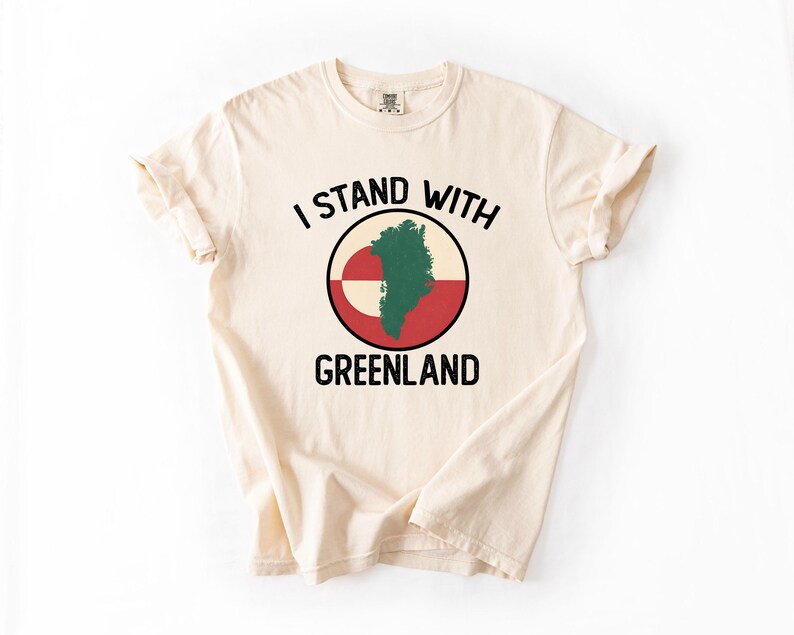 Pu&ograve; includere: T-shirt color crema con la scritta nera "I STAND WITH GREENLAND". Il design include una grafica circolare con la mappa della Groenlandia in verde, su sfondo rosso e bianco.