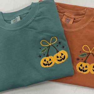 May include: Two Comfort Colors t-shirts, one teal and one rust-colored, are displayed. Each shirt features an embroidered design of two yellow jack-o'-lanterns with black faces, connected by a stem and leaves. The design is centered on the chest.