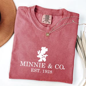 Pu&ograve; includere: Una maglietta colorata di rosso con la scritta "Minnie & Co. EST. 1928" e una grafica bianca di Minnie Mouse. Sono visibili anche una collana d'oro e un cappello marrone. La maglietta &egrave; piegata e posizionata su una superficie bianca.