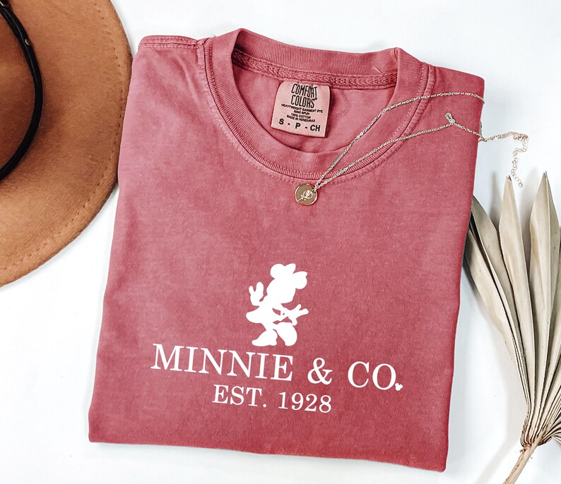 Pu&ograve; includere: Una maglietta colorata di rosso con la scritta "Minnie & Co. EST. 1928" e una grafica bianca di Minnie Mouse. Sono visibili anche una collana d'oro e un cappello marrone. La maglietta &egrave; piegata e posizionata su una superficie bianca.