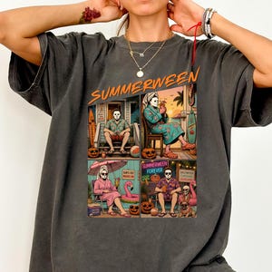 May include: A charcoal gray short-sleeve t-shirt featuring a graphic print with the text "SUMMERWEEN" at the top. The design consists of four panels depicting a masked character in various summer vacation settings with pumpkins and beach themes.