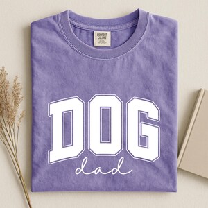 May include: A lilac-colored t-shirt with the words "DOG dad" printed in white. The word "DOG" is in a bold, collegiate-style font, and "dad" is in a cursive script. The shirt is folded neatly.