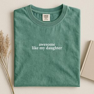 Puede incluir: Una camiseta verde agua doblada con el texto blanco "awesome like my daughter". La camiseta tiene cuello redondo y una etiqueta que dice "Comfort Colors". La camiseta est&aacute; sobre un fondo neutro.