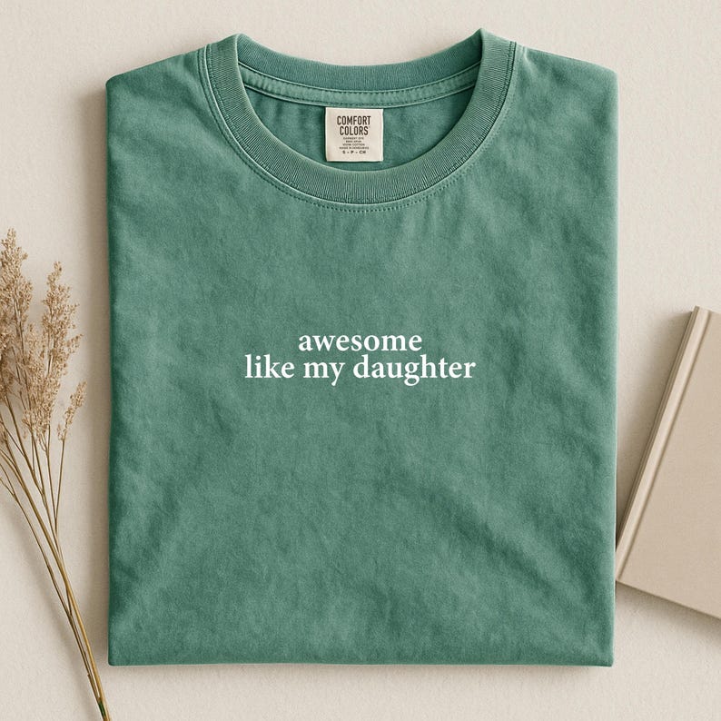 Puede incluir: Una camiseta verde agua doblada con el texto blanco "awesome like my daughter". La camiseta tiene cuello redondo y una etiqueta que dice "Comfort Colors". La camiseta est&aacute; sobre un fondo neutro.