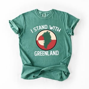 Pu&ograve; includere: T-shirt color verde acqua con la scritta bianca "I STAND WITH GREENLAND". La grafica presenta una mappa della Groenlandia verde all'interno di un cerchio rosso, bianco e nero. La maglietta &egrave; piegata.