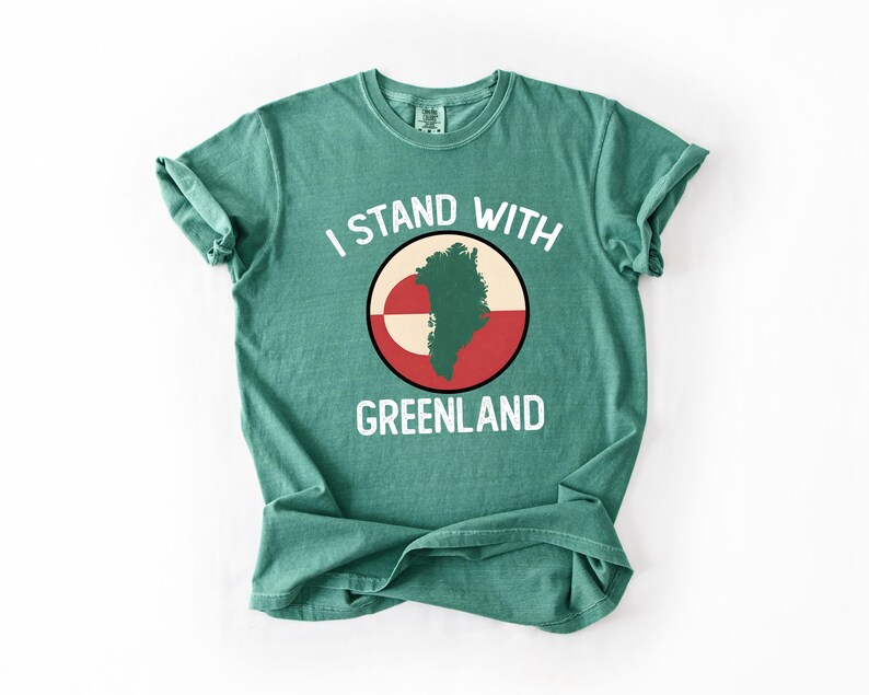 Pu&ograve; includere: T-shirt color verde acqua con la scritta bianca "I STAND WITH GREENLAND". La grafica presenta una mappa della Groenlandia verde all'interno di un cerchio rosso, bianco e nero. La maglietta &egrave; piegata.