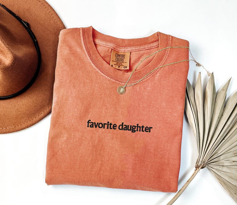 May include: A coral-coloured t-shirt with the phrase "favorite daughter" printed in black. A gold necklace with a pendant rests on the shirt. A brown hat and dried palm frond are also in the image.