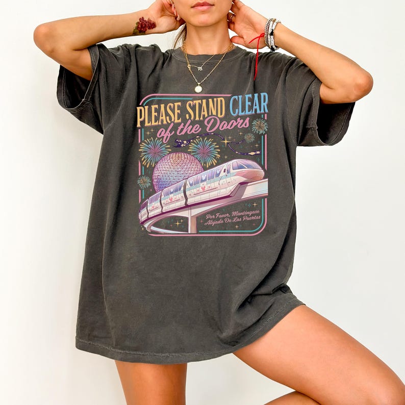 Puede incluir: Camiseta gris oscuro con un dise&ntilde;o gr&aacute;fico colorido que incluye un monorra&iacute;l, fuegos artificiales y una esfera, con el texto "PLEASE STAND CLEAR of the DOORS". La camiseta tiene mangas cortas y un corte holgado.