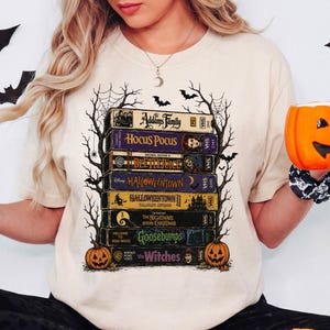 May include: A cream-colored t-shirt featuring a graphic print of a stack of VHS tapes titled The Addams Family, Hocus Pocus, Beetlejuice, Halloweentown, Halloweentown II, The Nightmare Before Christmas, Goosebumps, and The Witches, framed by spiderwebs.