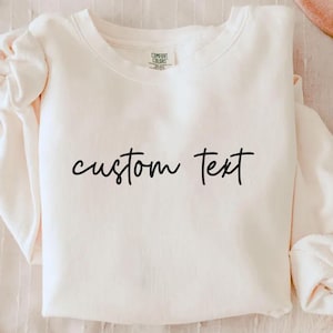 May include: Cream-colored sweatshirt with the words "custom text" in a handwritten style. The long-sleeved top is folded, with the sleeves bunched up. The tag "Comfort Colors" is visible on the neckline.