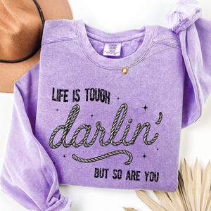 May include: A lavender crewneck sweatshirt with the text "Life is Tough Darlin' But So Are You" in black. The word "darlin'" is styled with a rope design. A gold necklace and a brown hat are also visible.