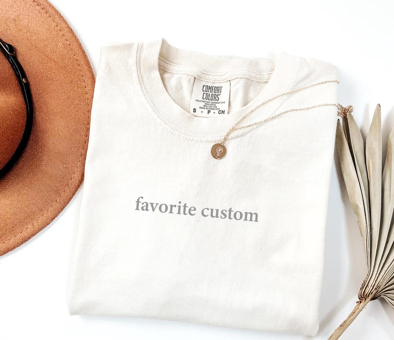K&ouml;nnte beinhalten: Cremefarbenes T-Shirt mit dem grauen Aufdruck "favorite custom". Eine goldene Halskette mit rundem Anh&auml;nger liegt auf dem Shirt. Ein brauner Hut und getrocknete Palmbl&auml;tter sind ebenfalls im Bild.
