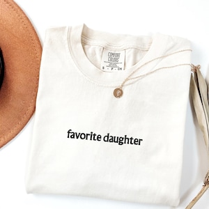 May include: Cream-coloured t-shirt with the embroidered text "favorite daughter" in black. A gold necklace with a circular pendant rests on the shirt. A brown hat and dried palm leaves are in the background.