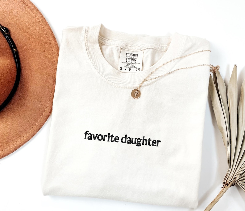 May include: Cream-coloured t-shirt with the embroidered text "favorite daughter" in black. A gold necklace with a circular pendant rests on the shirt. A brown hat and dried palm leaves are in the background.