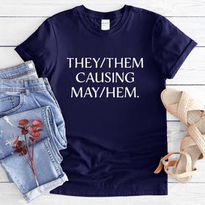May include: A navy blue short-sleeve t-shirt featuring the white text "THEY/THEM CAUSING MAY/HEM." alongside a pair of light blue denim jeans and a pair of beige woven sandals with ankle straps, arranged on a white wooden surface.