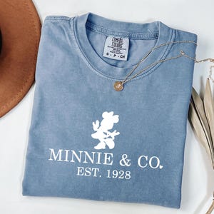 Pu&ograve; includere: Una maglietta blu con una grafica bianca di Minnie Mouse e la scritta "MINNIE & CO. EST. 1928". Una collana d'oro con un ciondolo &egrave; sulla maglietta. Un cappello marrone e una pianta secca sono sullo sfondo.