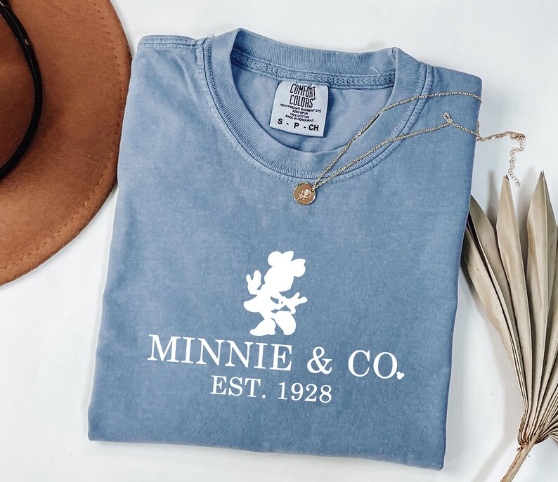 Pu&ograve; includere: Una maglietta blu con una grafica bianca di Minnie Mouse e la scritta "MINNIE & CO. EST. 1928". Una collana d'oro con un ciondolo &egrave; sulla maglietta. Un cappello marrone e una pianta secca sono sullo sfondo.