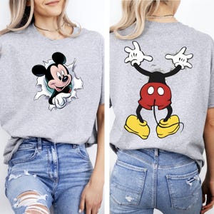 May include: A heather gray t-shirt featuring a graphic of Mickey Mouse peeking through a torn paper effect on the front, and the lower half of Mickey Mouse with red shorts and yellow shoes on the back, paired with blue denim jeans.