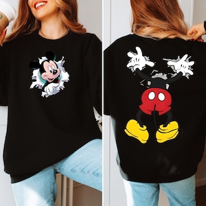 May include: Black sweatshirt featuring a Mickey Mouse graphic on the front and back. The front design shows Mickey breaking through a white-edged hole. The back displays Mickey's body, with red shorts, yellow shoes, and white gloves.