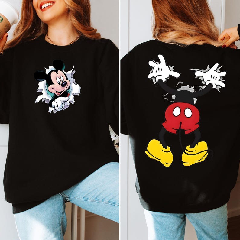 May include: Black sweatshirt featuring a Mickey Mouse graphic on the front and back. The front design shows Mickey breaking through a white-edged hole. The back displays Mickey's body, with red shorts, yellow shoes, and white gloves.