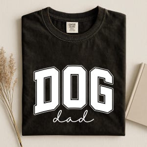 May include: Black t-shirt with the white text "DOG dad" in a bold, collegiate-style font. The shirt has a crew neck and a tag that says "Comfort Colors". The shirt is folded on a light-colored surface.