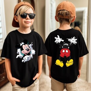 May include: A black t-shirt featuring a graphic of Mickey Mouse on the front and back. The front shows Mickey's head and hand emerging from a white torn paper effect. The back displays Mickey's lower body, red shorts, and yellow shoes.