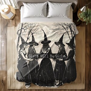 Peut inclure: Une couverture beige et noire avec une illustration vintage de cinq femmes portant des chapeaux et des robes de sorcières. Le texte "Girls Will Be Girls" est imprimé au centre. La couverture est sur un lit avec des oreillers blancs.
