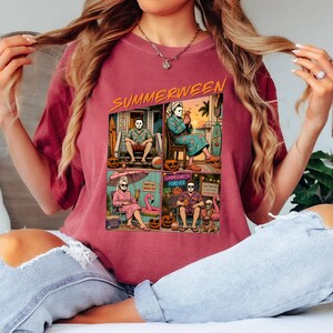 May include: A maroon t-shirt featuring a graphic print with the text "SUMMERWEEN" at the top. The graphic is divided into four panels showing a masked character in various summer settings, including a porch, a patio, a beach chair, and a lounge.