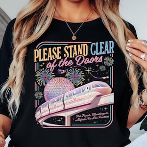 Puede incluir: Camiseta negra con un gr&aacute;fico colorido que presenta un monorra&iacute;l, fuegos artificiales y una esfera geod&eacute;sica. El texto dice "PLEASE STAND CLEAR of the Doors" y "Por Favor, Mant&eacute;ngase Alejado De Las Puertas."