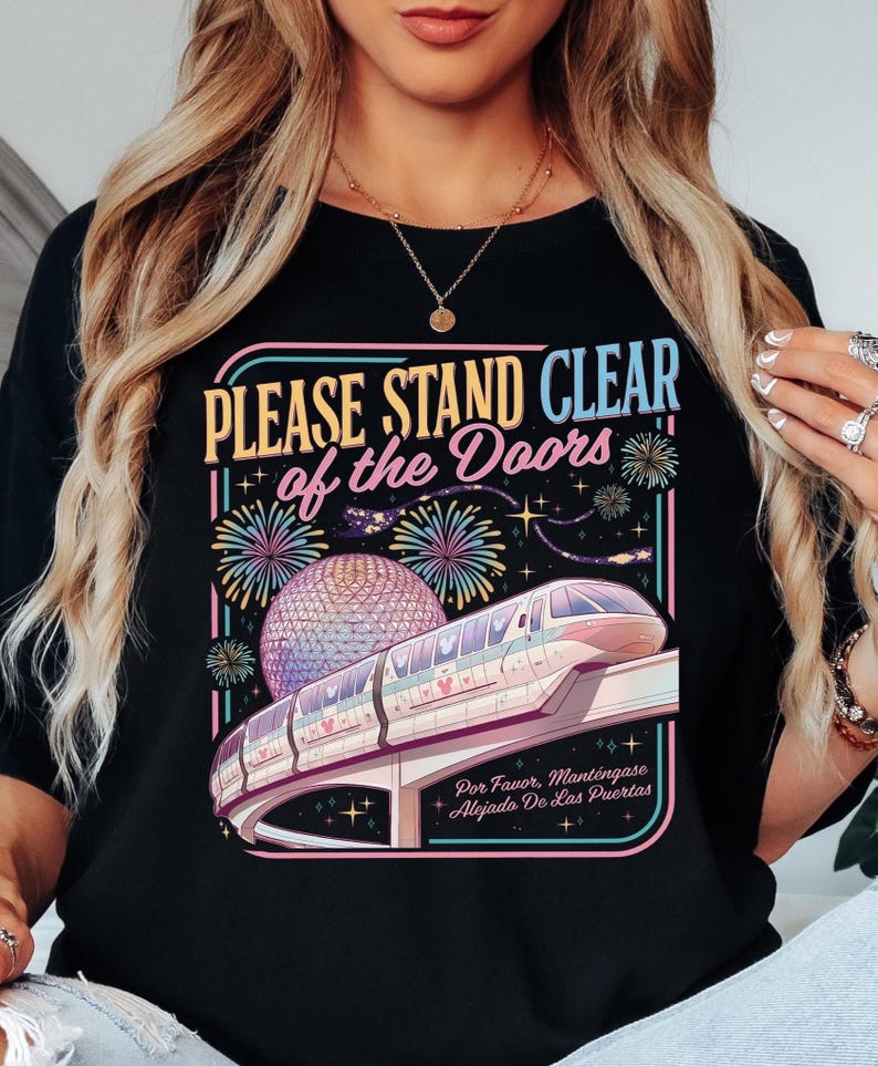 Puede incluir: Camiseta negra con un gr&aacute;fico colorido que presenta un monorra&iacute;l, fuegos artificiales y una esfera geod&eacute;sica. El texto dice "PLEASE STAND CLEAR of the Doors" y "Por Favor, Mant&eacute;ngase Alejado De Las Puertas."