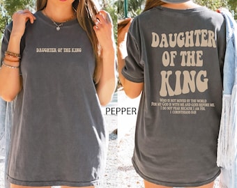 May include: A pepper-colored t-shirt featuring the text DAUGHTER OF THE KING on the front. The back displays the same text in large, wavy beige letters, followed by the verse: WHO IS NOT MOVED BY THE WORLD FOR MY GOD IS WITH ME AND GOES BEFORE ME. I DO NOT FEAR BECAUSE I AM HIS. 1 CORINTHIANS 6:18.