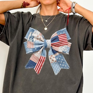 May include: Dark grey t-shirt with a large bow design. The bow is made of patchwork denim and fabric with American flag and floral patterns. The bow has a white lace centre and frayed edges.