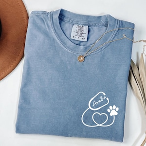 May include: A blue Comfort Colors t-shirt with a white stethoscope design, a heart, a paw print, and the name "Amelia". A gold necklace with a pendant is on the shirt. The shirt is folded on a white surface.