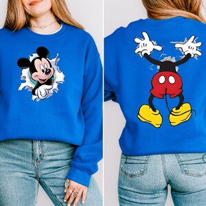 May include: A royal blue sweatshirt featuring Mickey Mouse. The front shows Mickey popping out of a torn hole, while the back displays Mickey's full body with outstretched arms. The cartoon character is in his classic red shorts and yellow shoes.