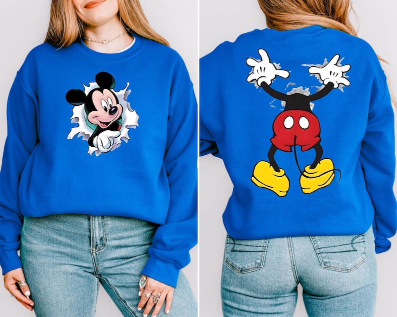 May include: A royal blue sweatshirt featuring Mickey Mouse. The front shows Mickey popping out of a torn hole, while the back displays Mickey's full body with outstretched arms. The cartoon character is in his classic red shorts and yellow shoes.